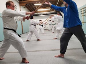 people training in Aikido. Most are wearing traditional white cotten dogi, but one person is wearing sweat pants and a long-sleeved knit shirt. Everyone's knees and elbows are covered.